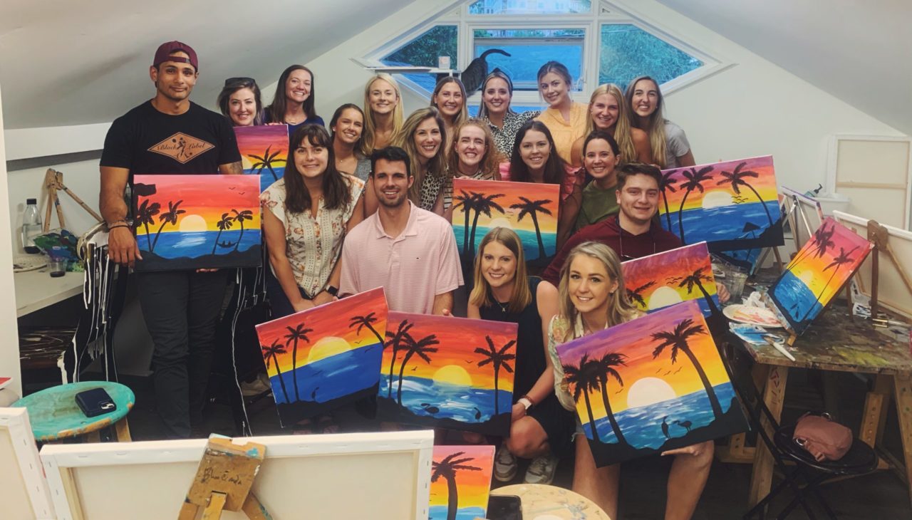 Group of people holding paintings of a tropical sunset