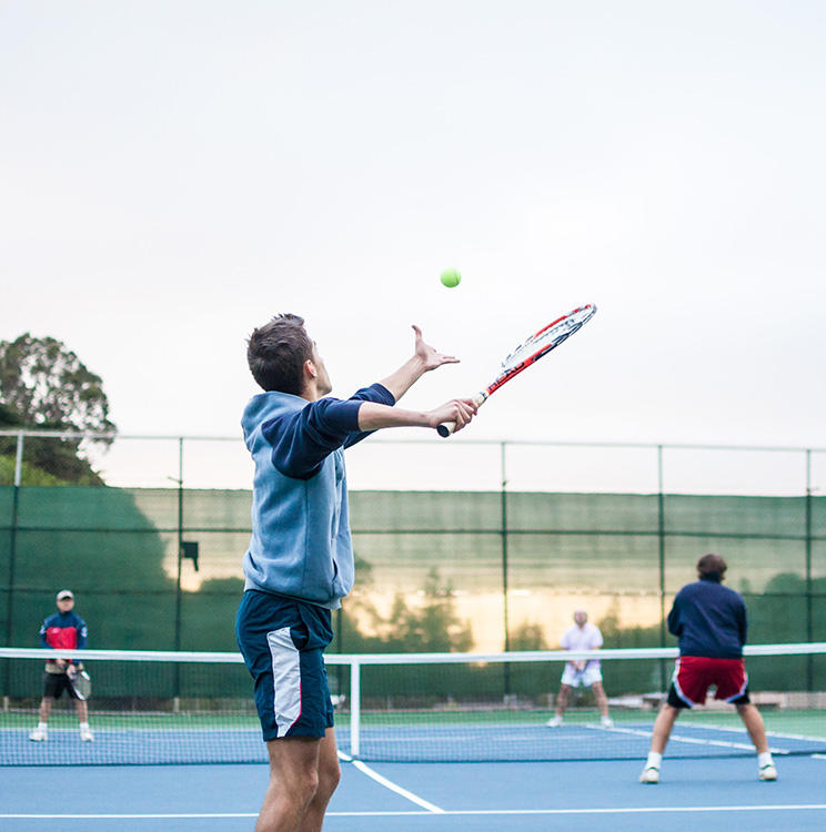 A man serves a tennis ball during a doubles tennis match.