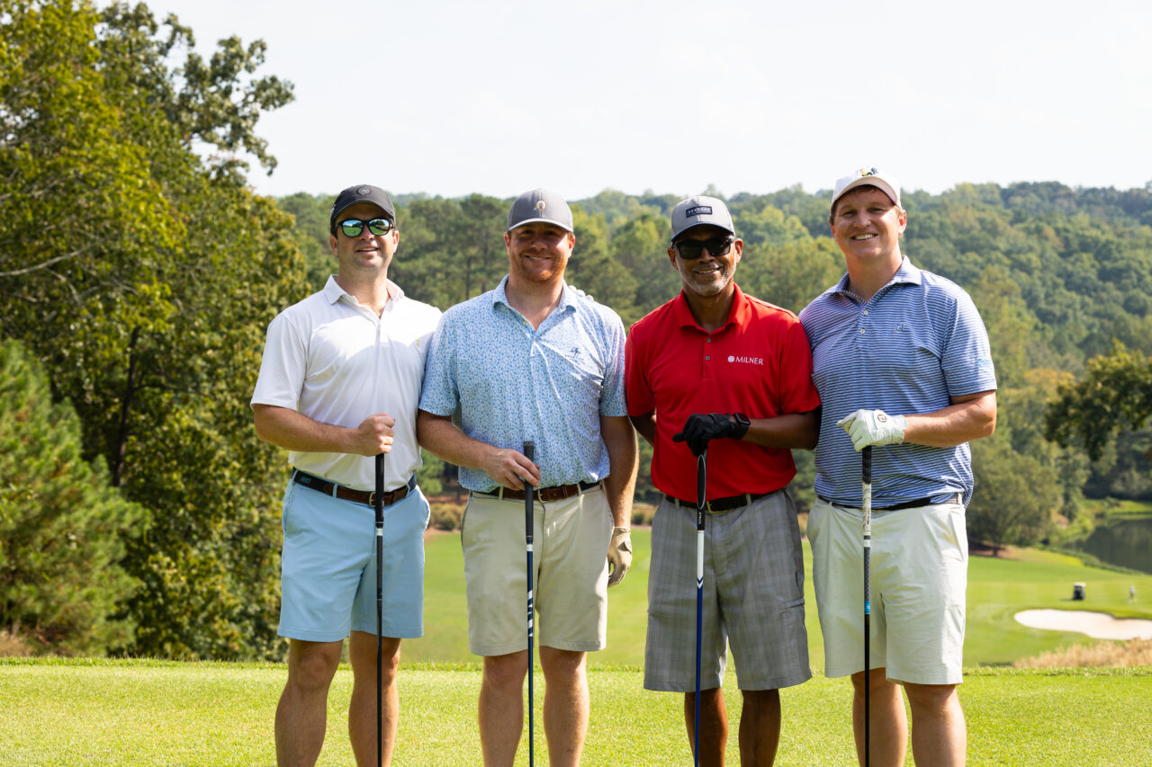 Four men smile while they stand on a golf course and hold golf clubs