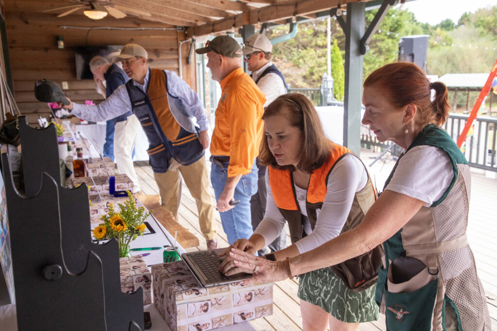 A small group of people in outdoor gear engages in an activity on a wooden deck. Two women focus on a laptop while three men observe a lineup of items on tables in the background.