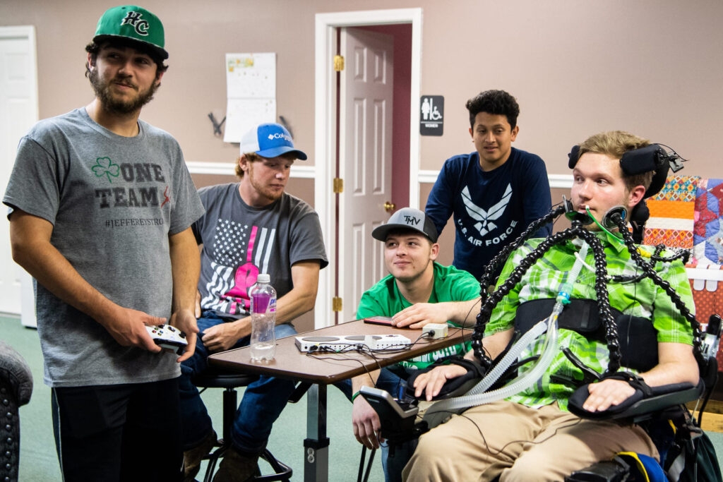 A group of five people, one in a motorized wheelchair, is gathered around a table with gaming consoles and screens in a room.