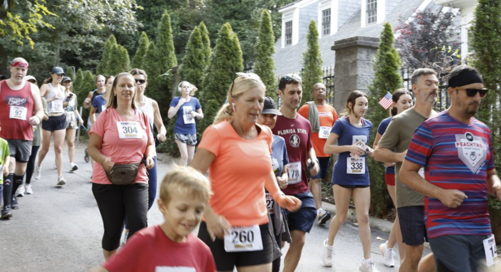 A group of people, including adults and children, participate in an outdoor race, jogging down a road lined with trees. Many wear numbered race bibs.