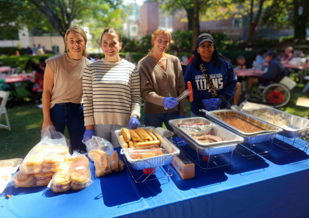 Four women stand behind a blue table outdoors, serving hot dogs and other food items from metal trays at an event. People are seated at tables in the background.