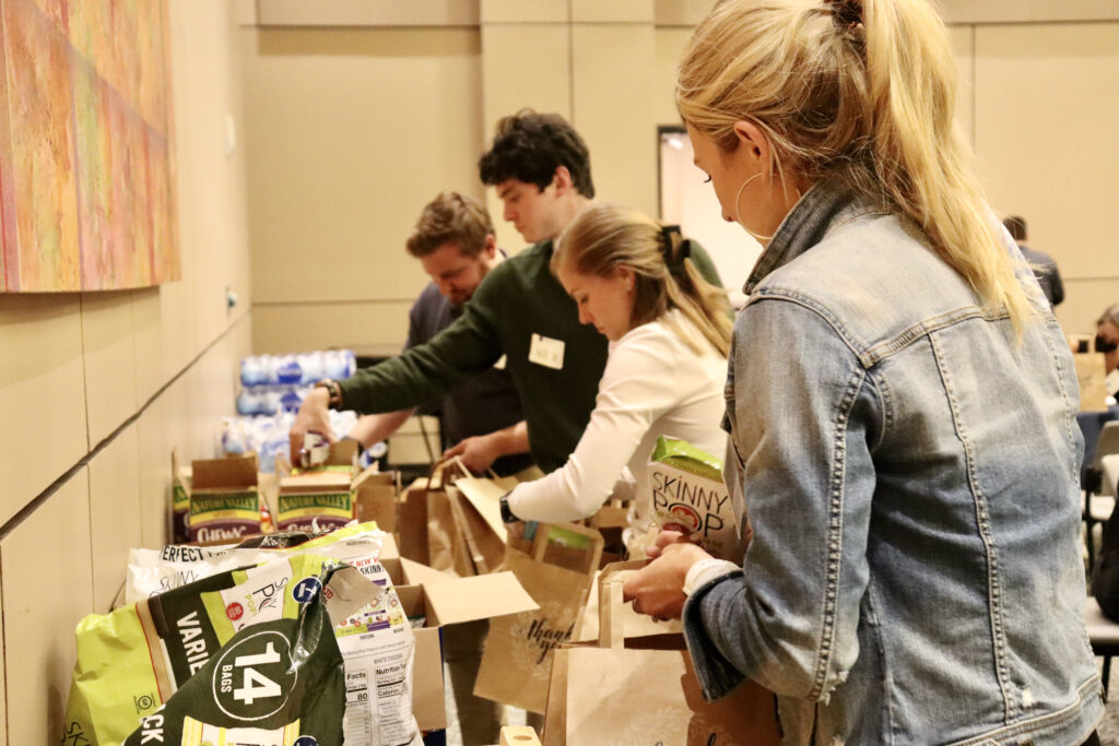Four people are packing groceries into paper bags at a table.