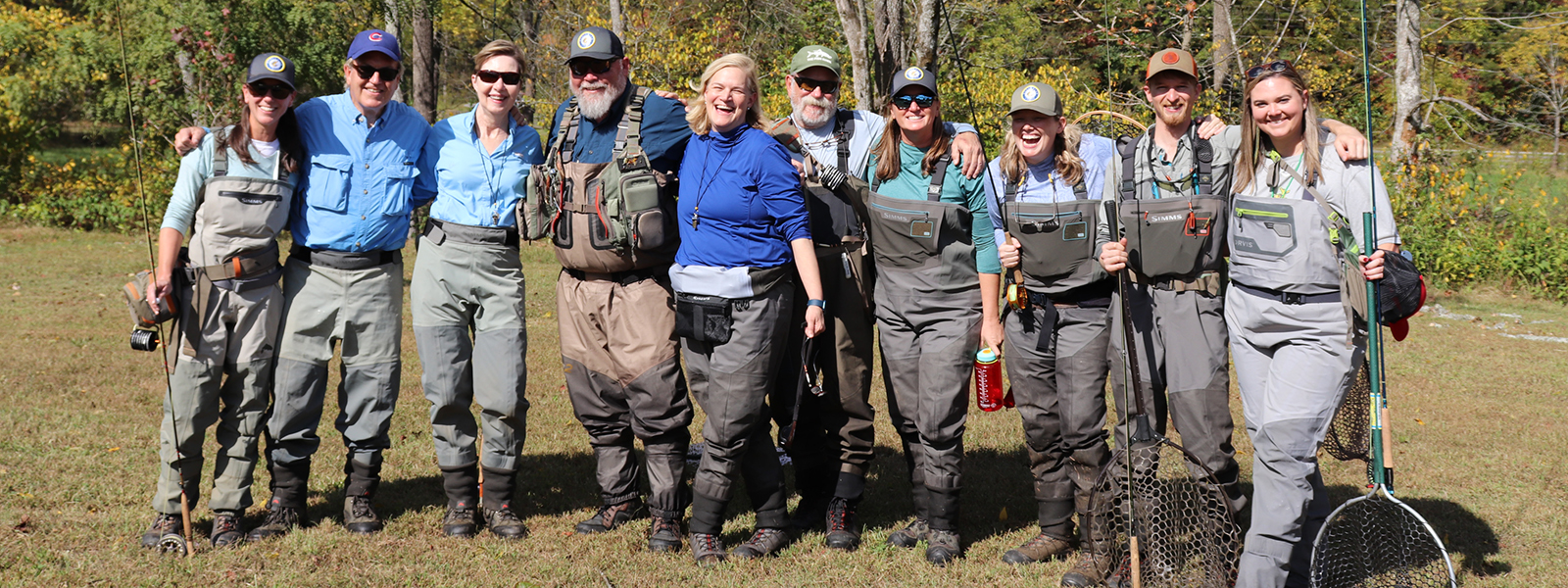 A group of ten people wearing fishing gear and waders stand outdoors in a line, smiling at the camera.