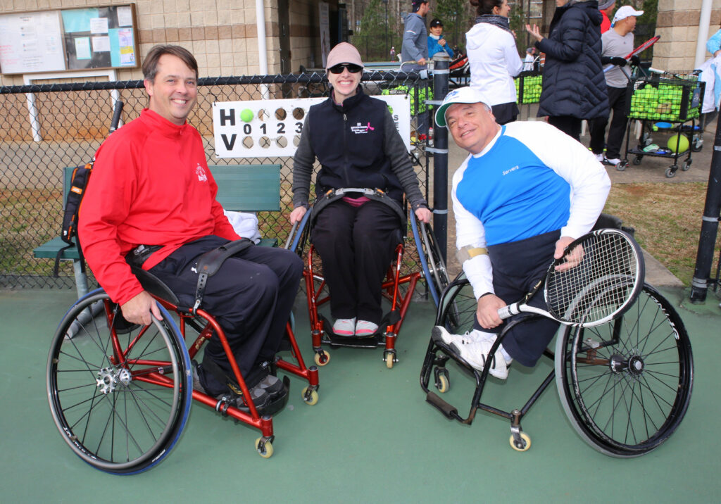 Three tennis players in wheelchairs on a tennis court, with other players and equipment in the background.