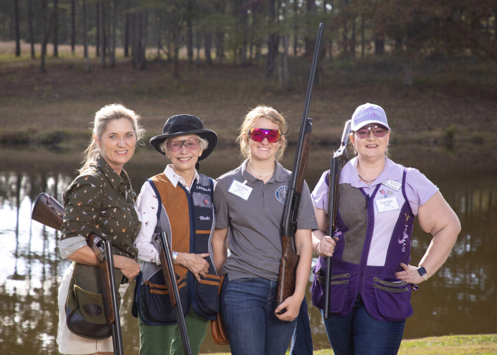 Four women holding rifles stand together outdoors near a pond, dressed in shooting gear.