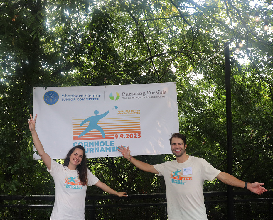 Two people stand in front of a banner for a Cornhole Tournament held by the Shepherd Center, raising their arms in excitement.