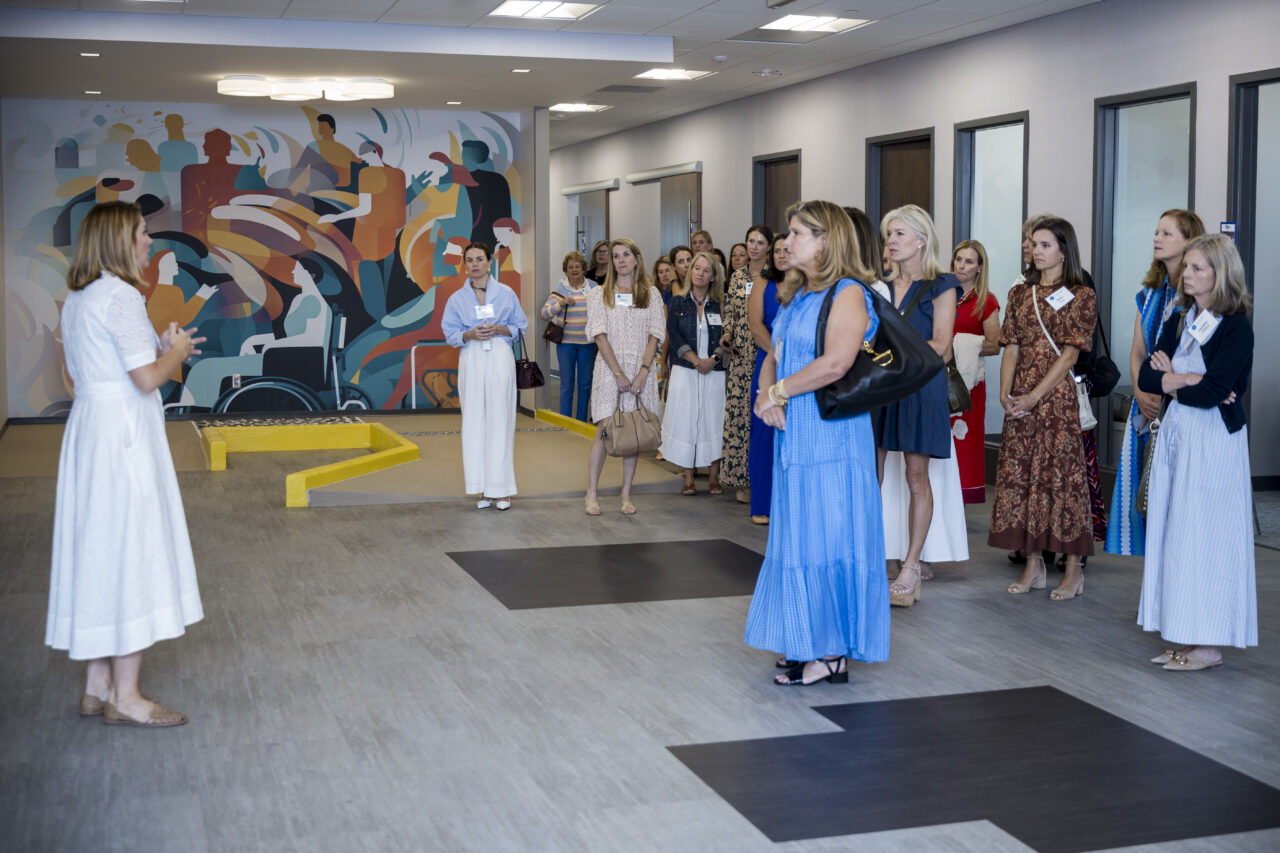 A group of women stand and listen to a woman speaking in a modern room with a colorful mural.