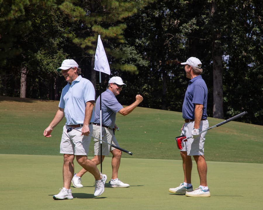 Three men on a golf course celebrate a successful putt. One man pumps his fist in victory, while the others smile.